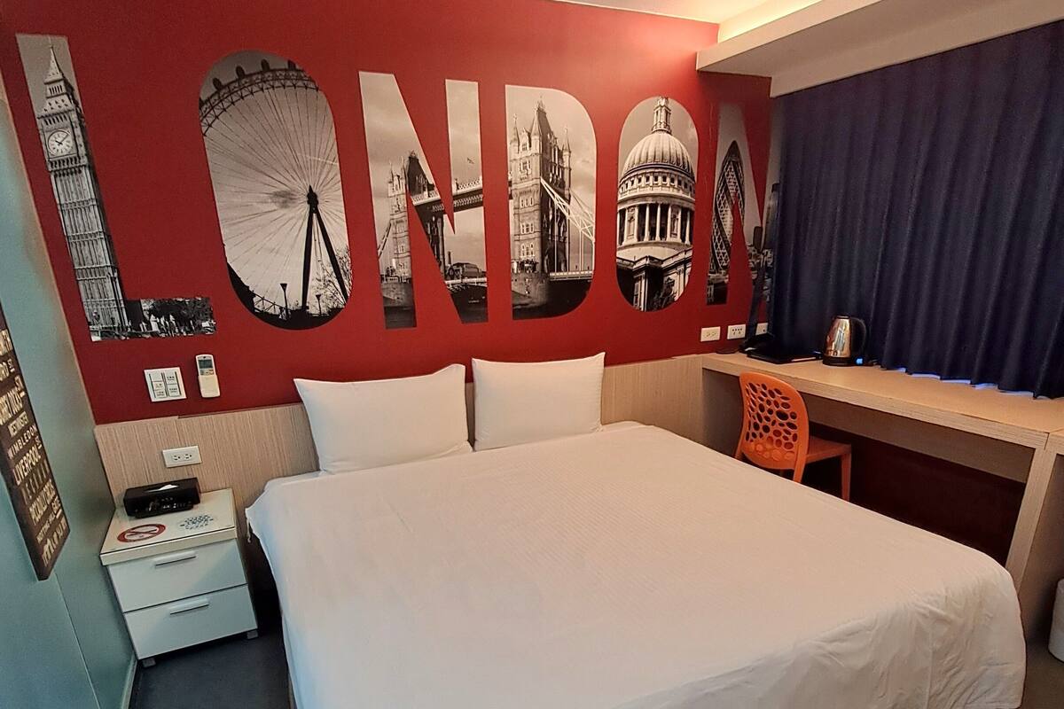 Une chambre d’hôtel avec un grand lit, un bureau et une chaise. Un mur est décoré du mot « LONDRES » et d’images de monuments emblématiques.