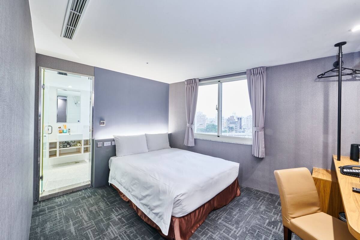 A hotel room with a bed, a desk, a chair, a window with curtains, and a view of the city.