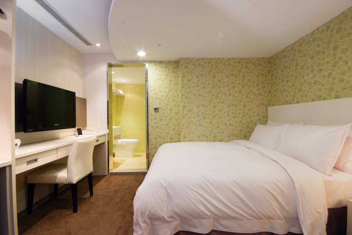 A hotel room with a bed, a desk with a flat-screen TV, a chair, and a bathroom visible through an open door.