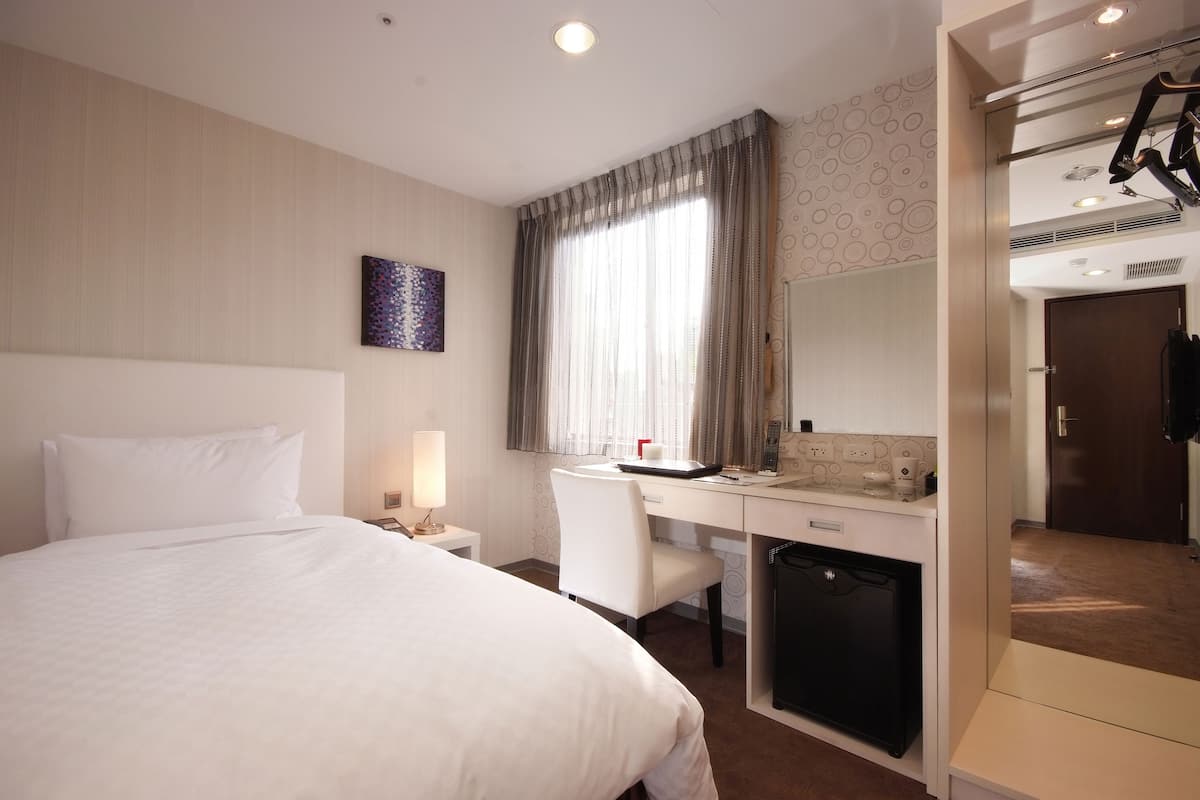 A hotel room with a bed, desk, chair, and a small kitchenette.