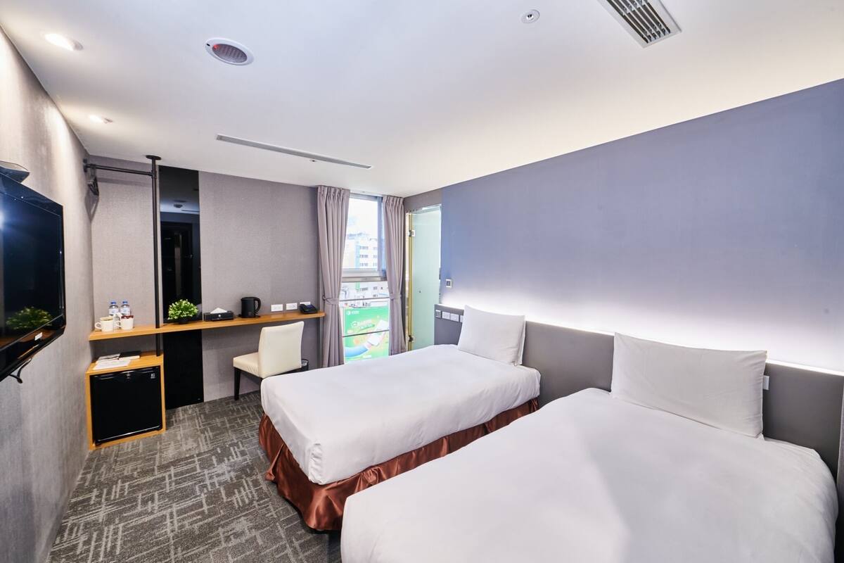 A hotel room with two beds, a desk with a chair, a small fridge, and a TV.