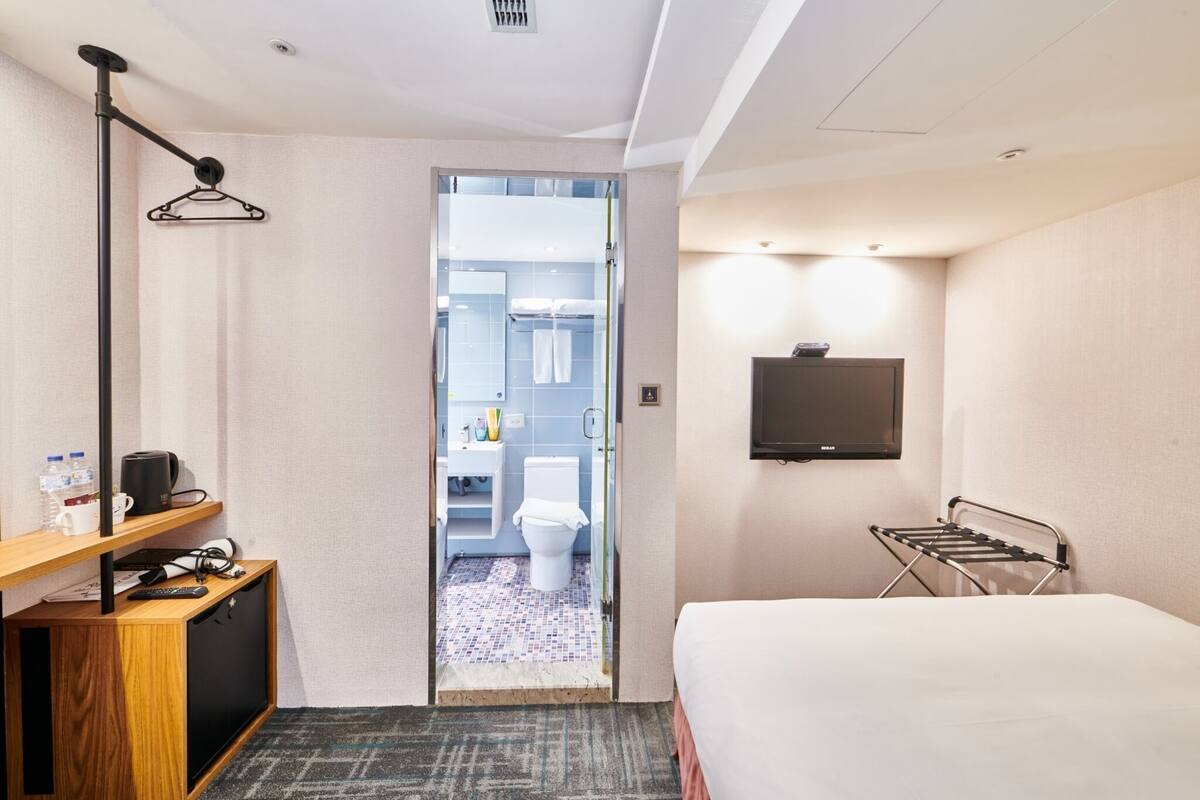 A hotel room with a bed, a TV mounted on the wall, a desk with a lamp, a hanging clothes rack, and a view into the bathroom.