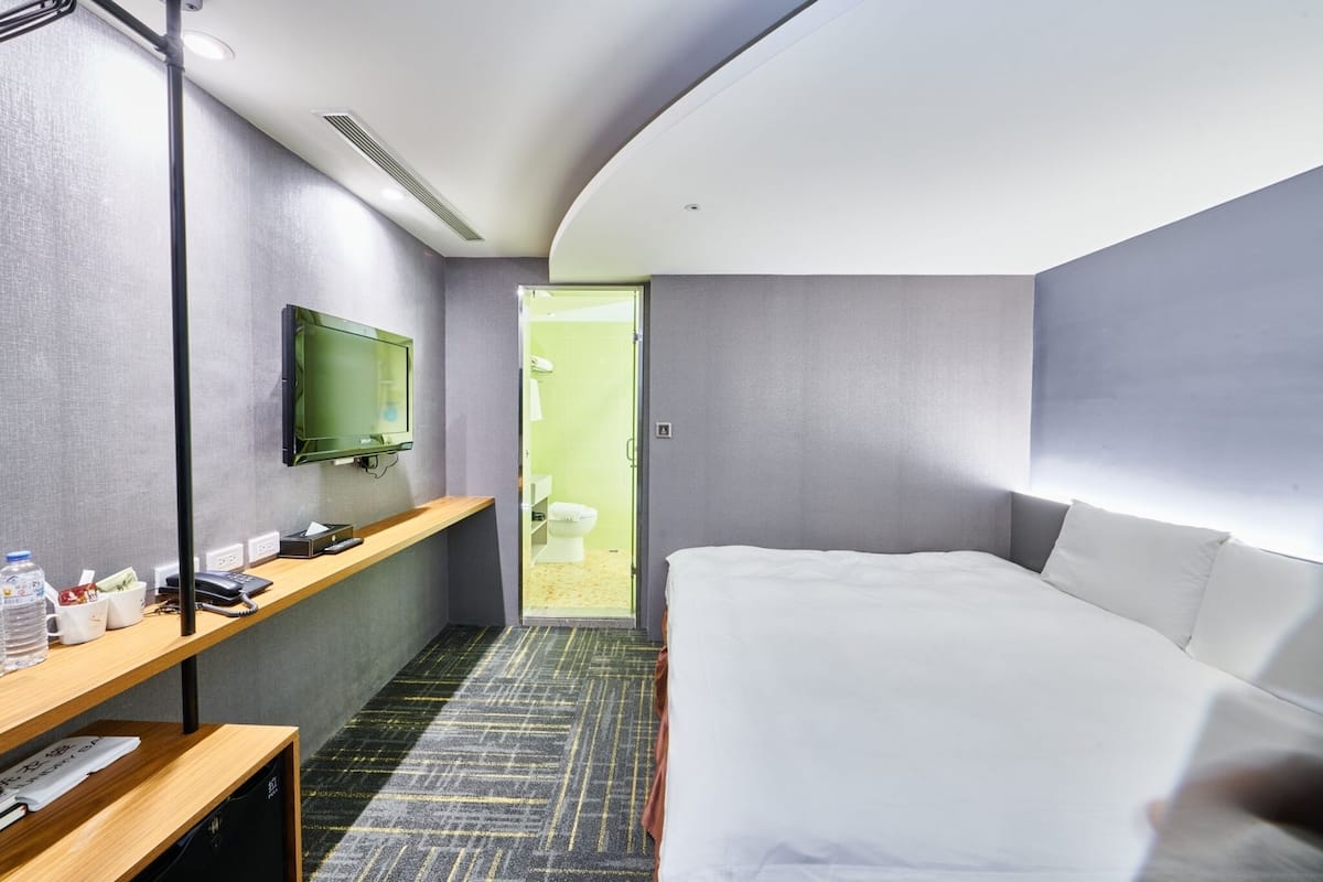 A hotel room with a bed, a desk with a flat-screen TV, a shelf with toiletries, and a door leading to a bathroom.