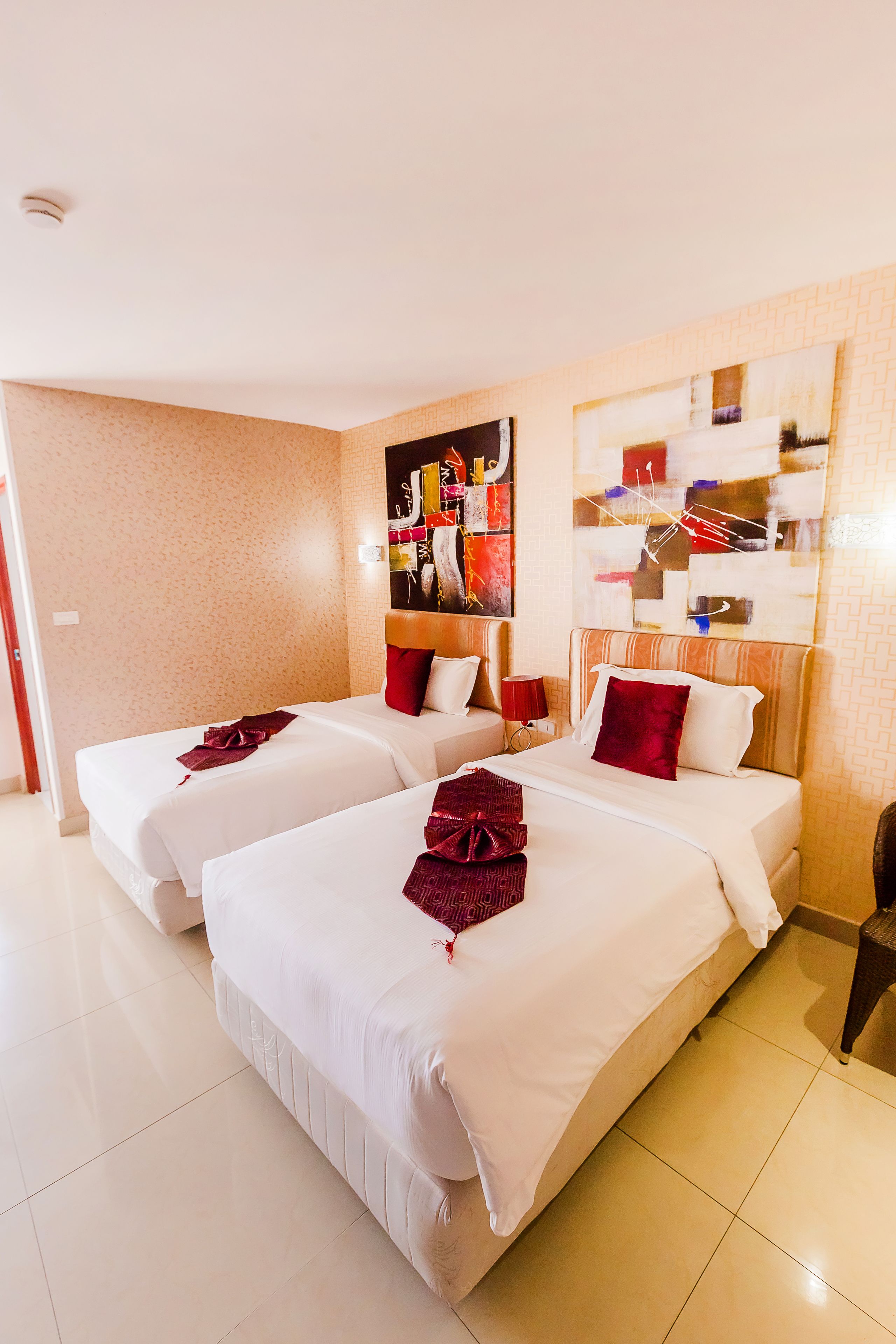 A hotel room with two beds, a red throw on each bed, a wall art piece, and a small table with a lamp.
