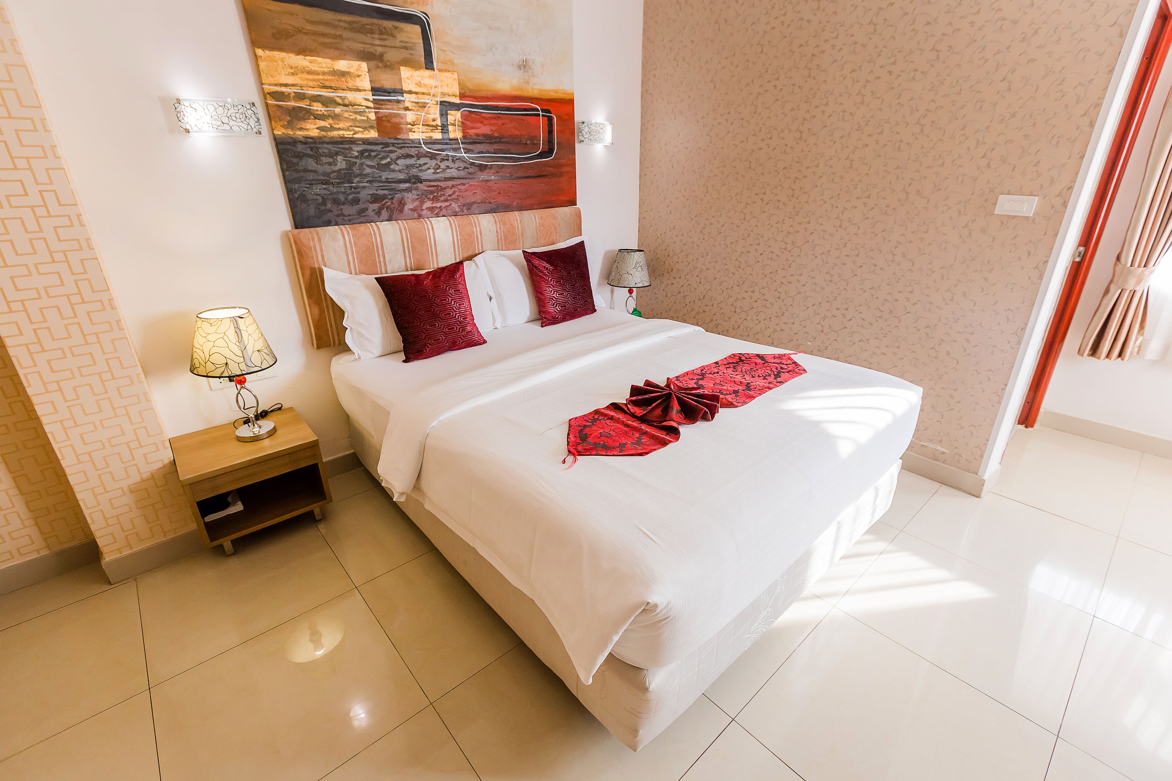 A neatly made bed with white linens and red accents, a bedside table with a lamp, and a large abstract painting above the bed.