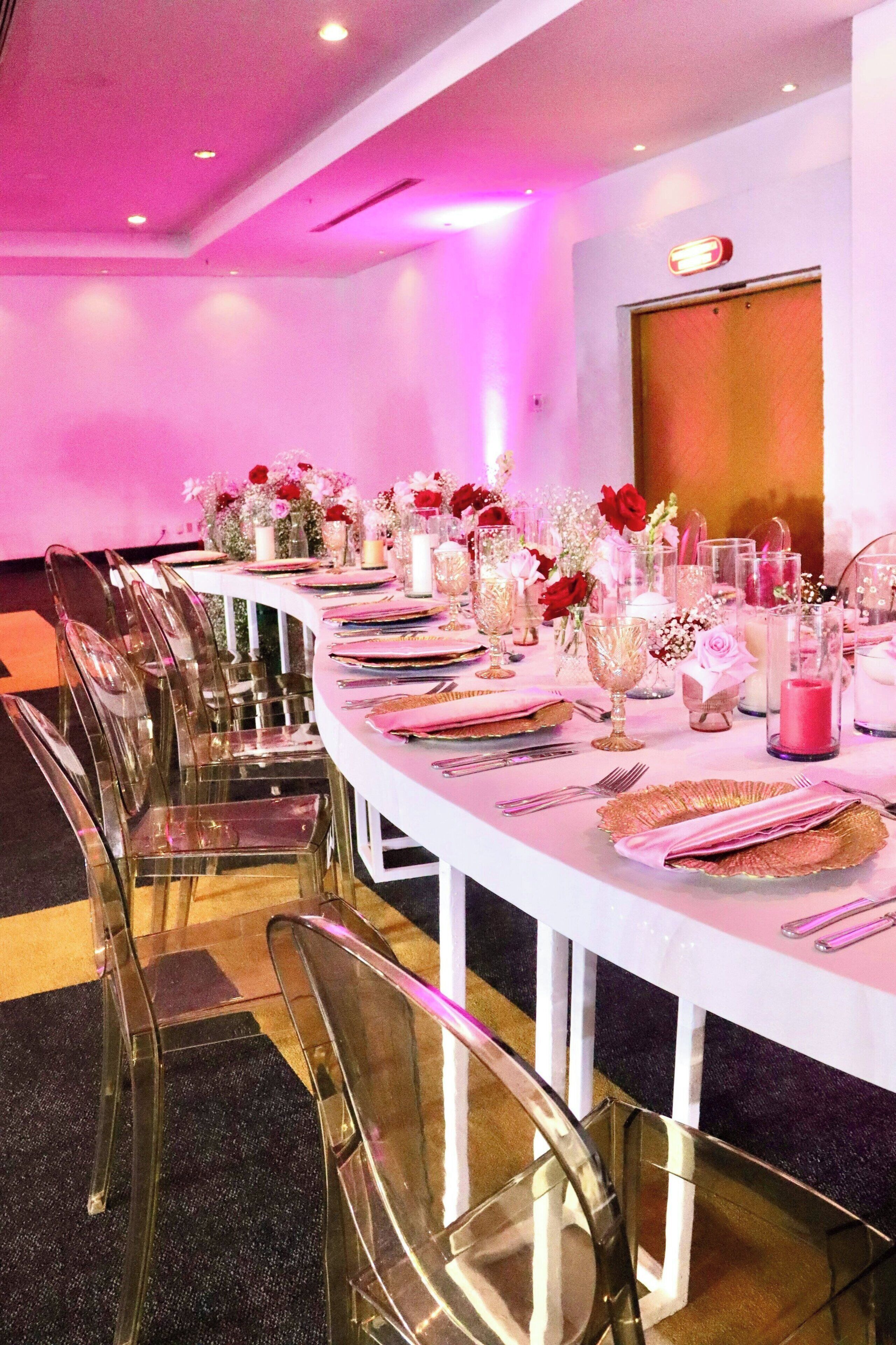 Una larga mesa dispuesta para un evento formal, con sillas traslúcidas, centros de mesa florales y vajilla elegante.