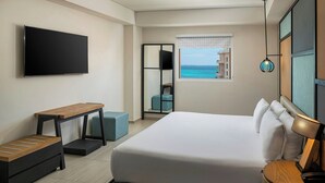 A modern hotel room with a large bed, a flat-screen TV mounted on the wall, a wooden bench, a desk, and a view of the ocean through a window.