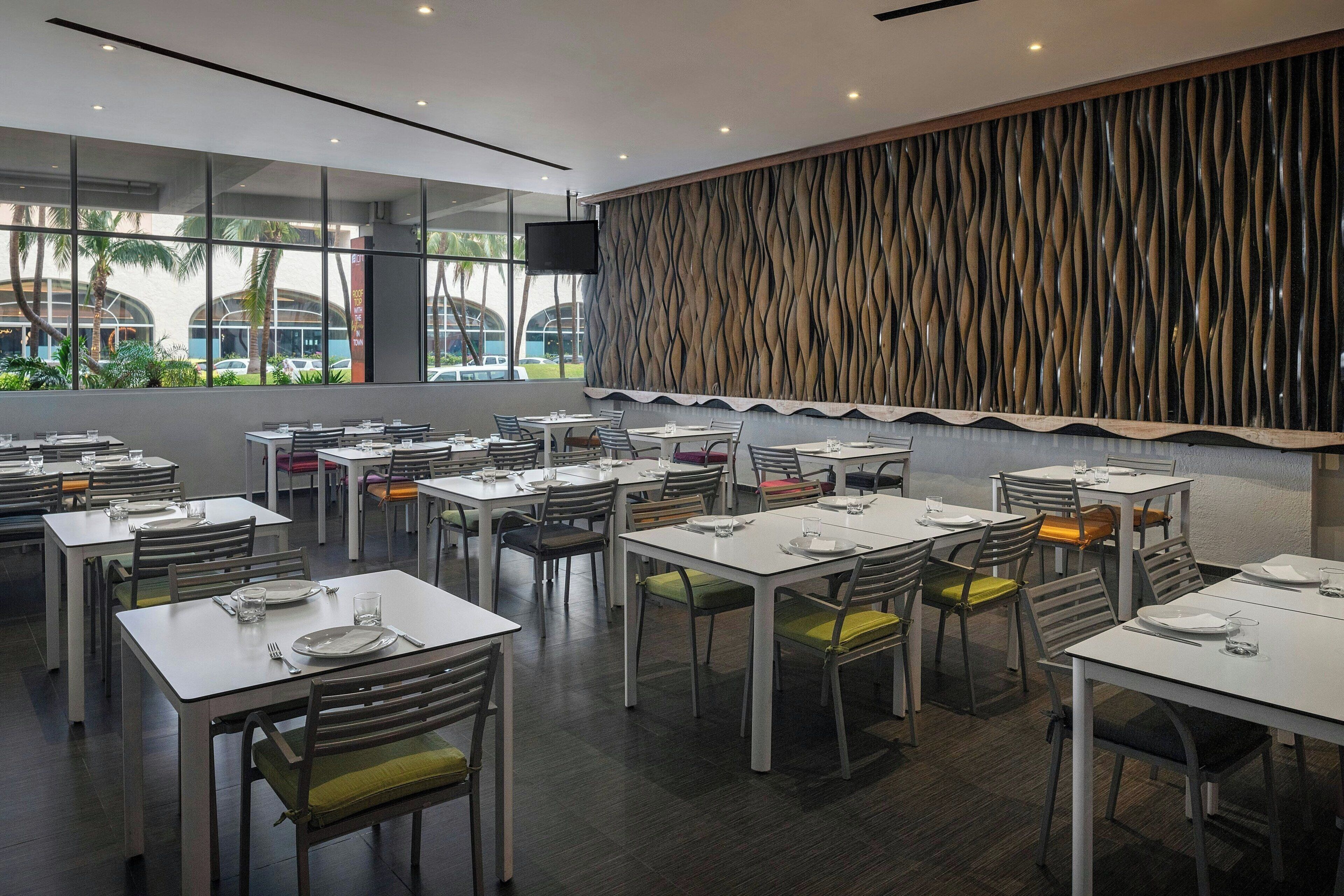 A modern restaurant with white tables and chairs, large windows, and a decorative wall.