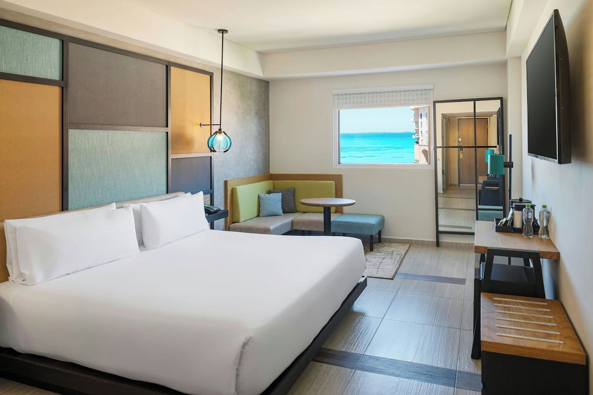 Habitación de hotel con una cama grande, zona de estar con vista al mar, una mesita y televisor montado en la pared.