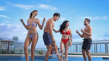 Un grup de persones gaudeix d'una piscina sota un cel blau clar amb edificis al fons.
