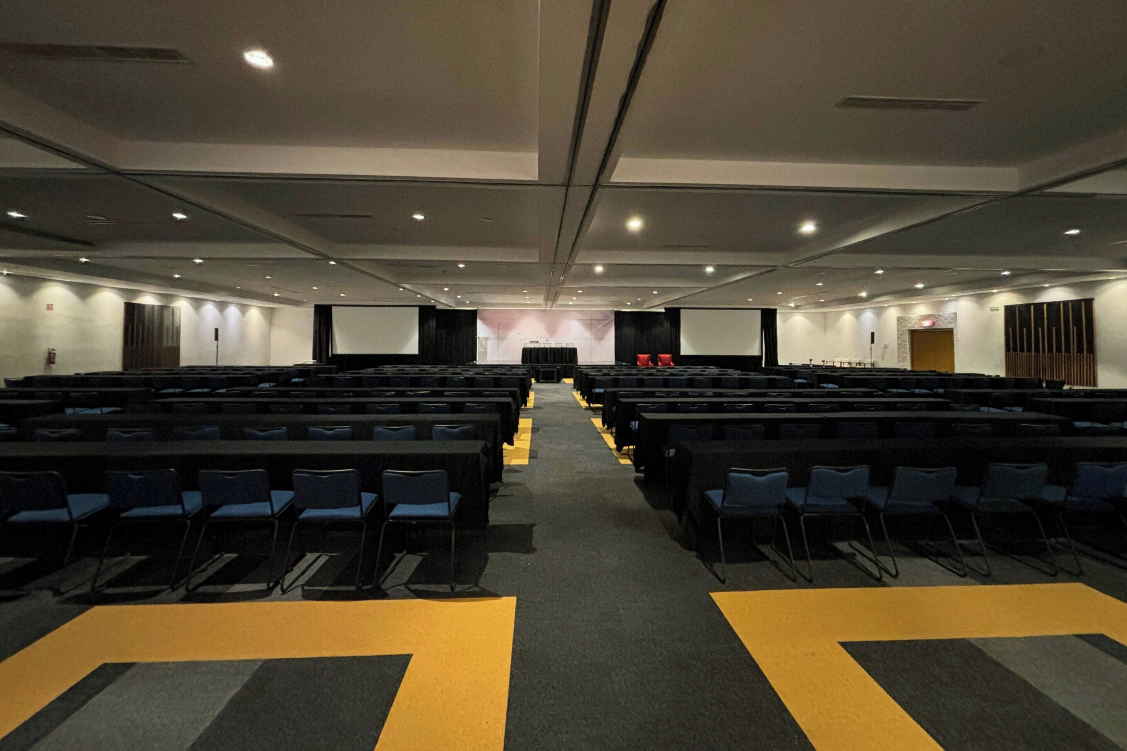 Un salón de conferencias con filas de sillas, un escenario al frente y una pantalla de proyector.
