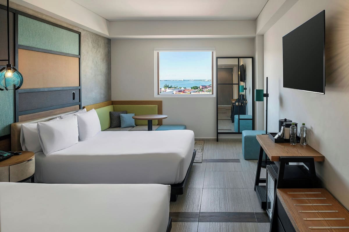 Habitación de hotel con dos camas, una mesita, un sofá, un televisor y una ventana con vistas al mar.