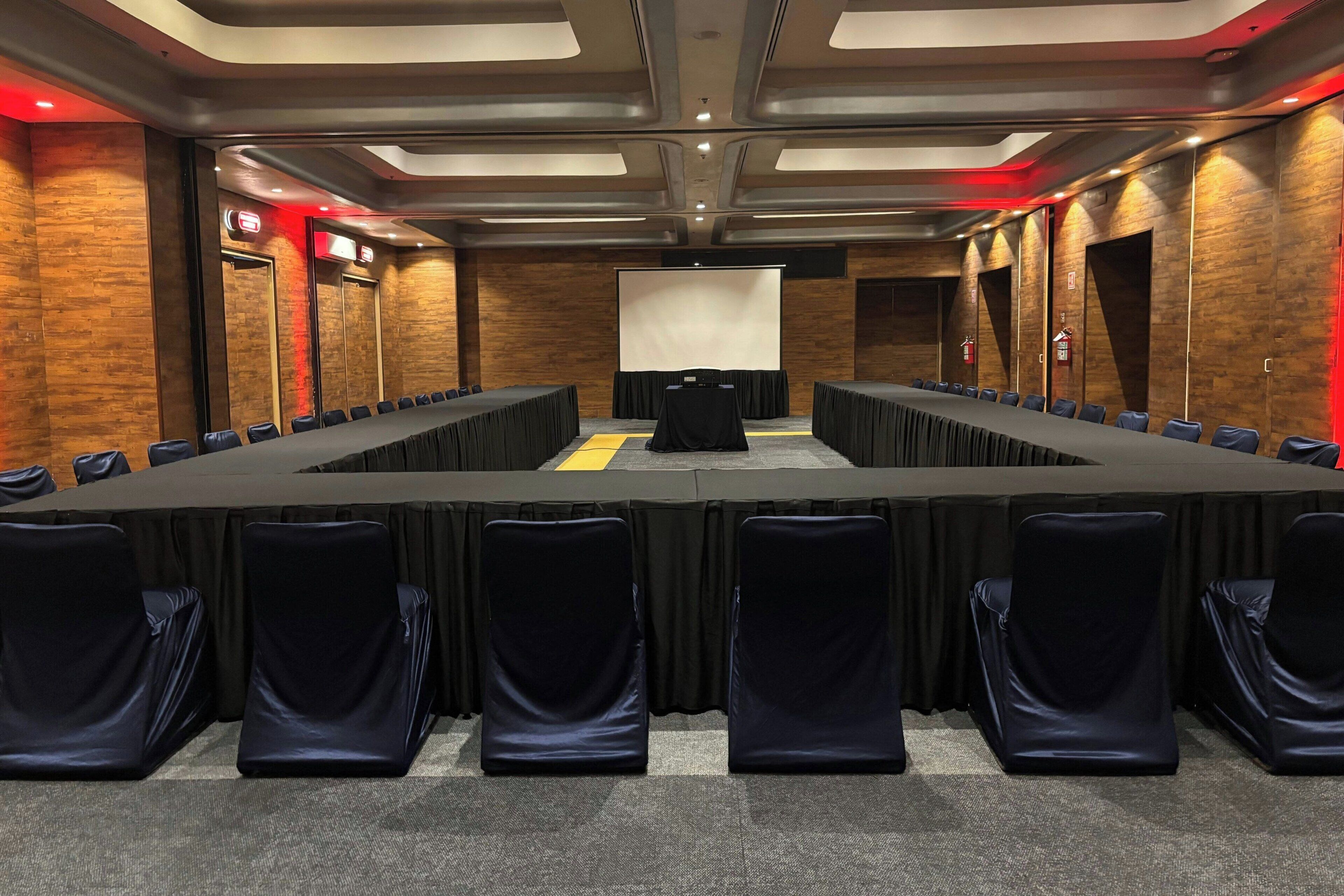 A conference room with a long table, chairs, a projector screen, and wooden walls.