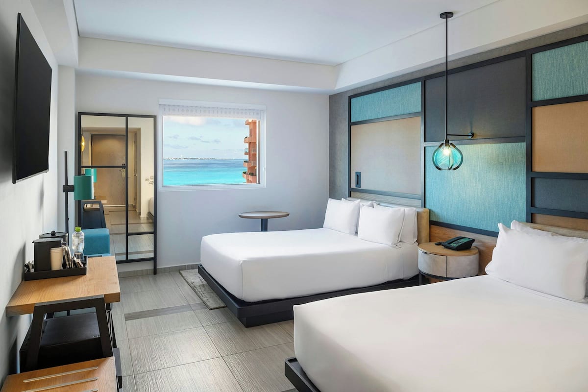 Habitación de hotel con dos camas, un escritorio, una silla, una mesita y una ventana con vistas al mar.