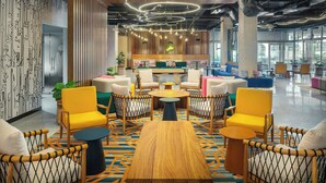 A modern lounge area with a central wooden table, surrounded by yellow and white chairs, and a patterned carpet.