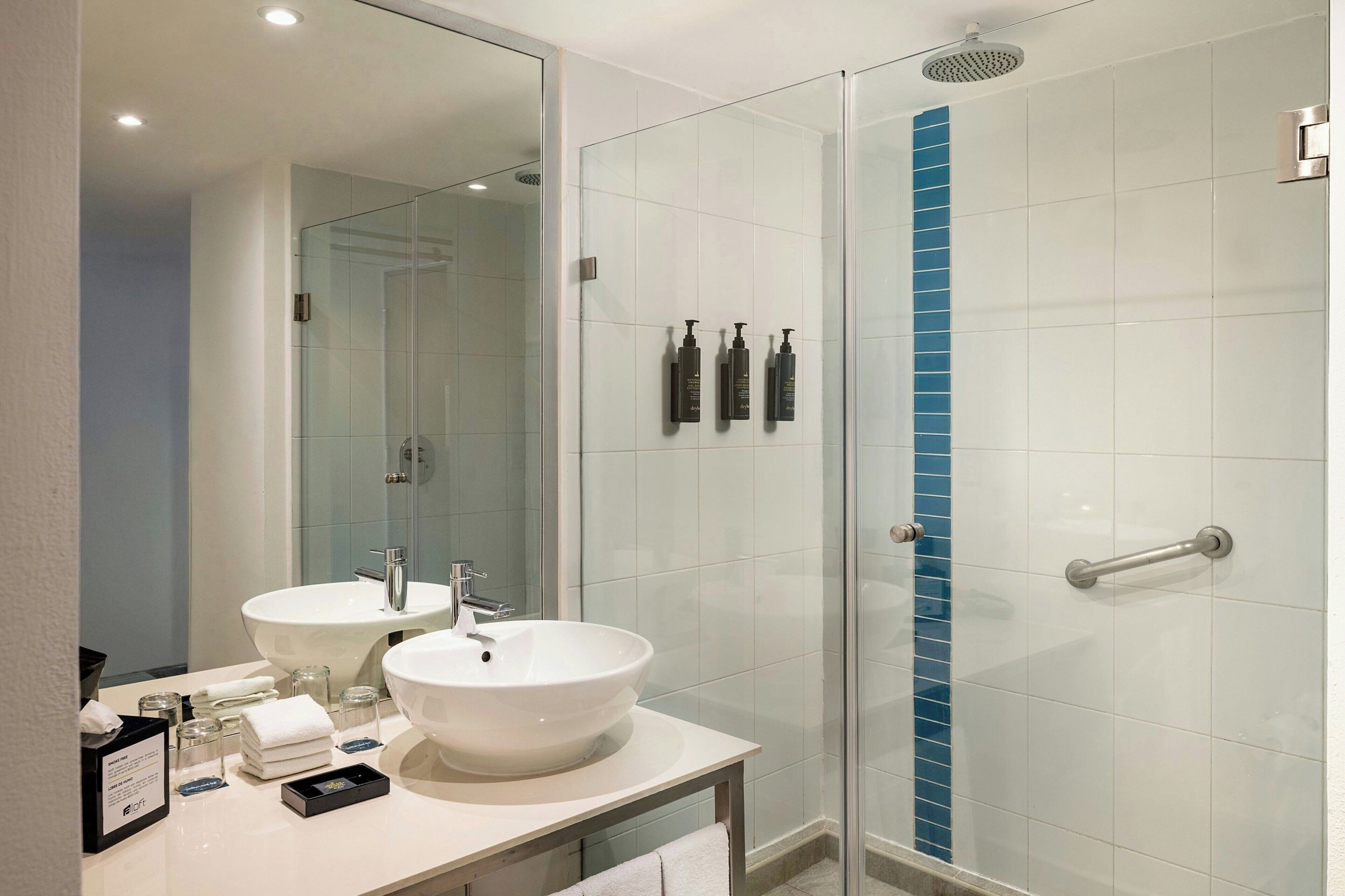 A modern bathroom with two sinks, a glass shower enclosure, and toiletries on the counter.