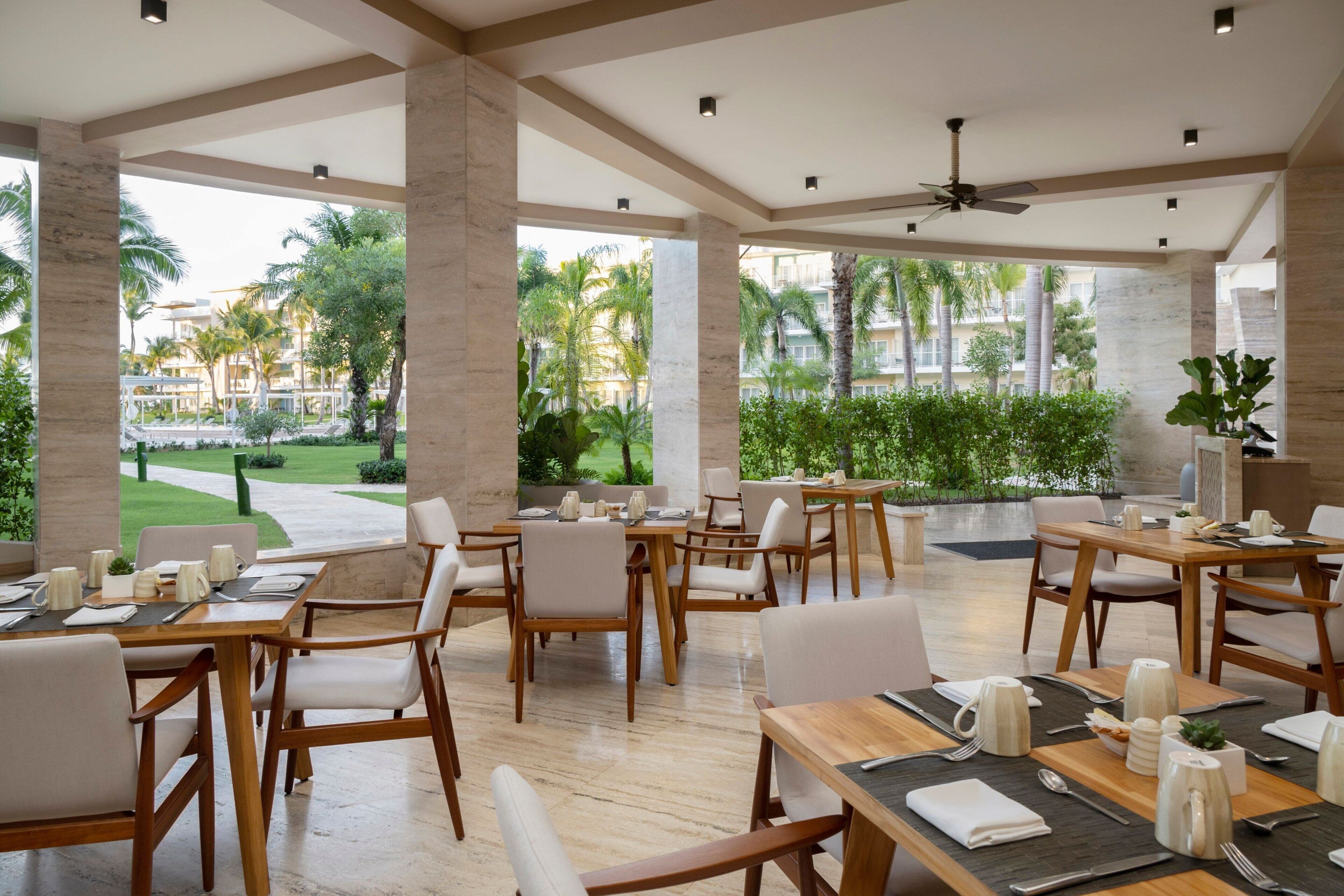 Un'ampia zona pranzo all'aperto con tavoli e sedie, e grandi finestre che offrono vista su un giardino curato e edifici.
