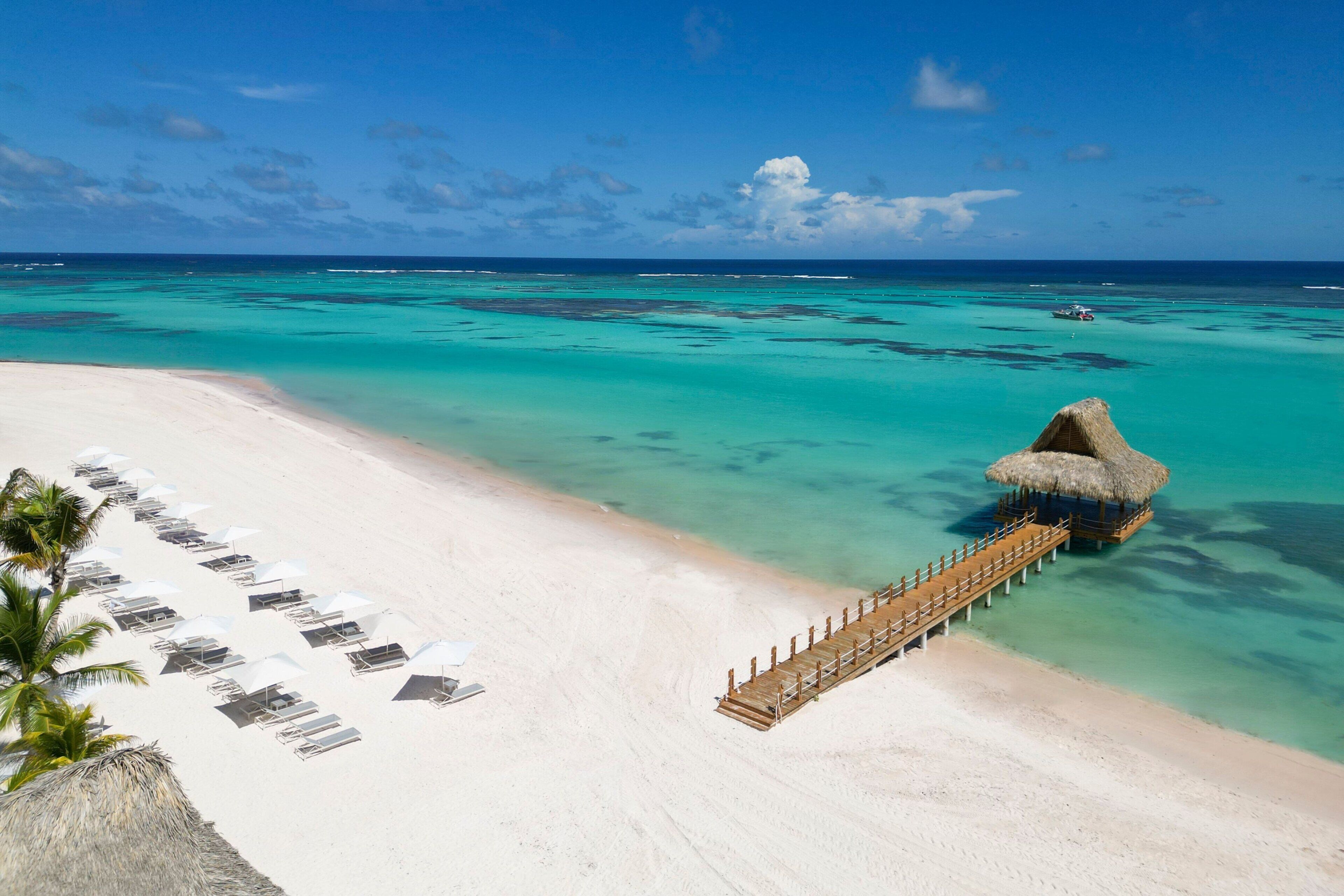 A beach with white sand, clear turquoise water, and a wooden pier leading to a thatched-roof structure.