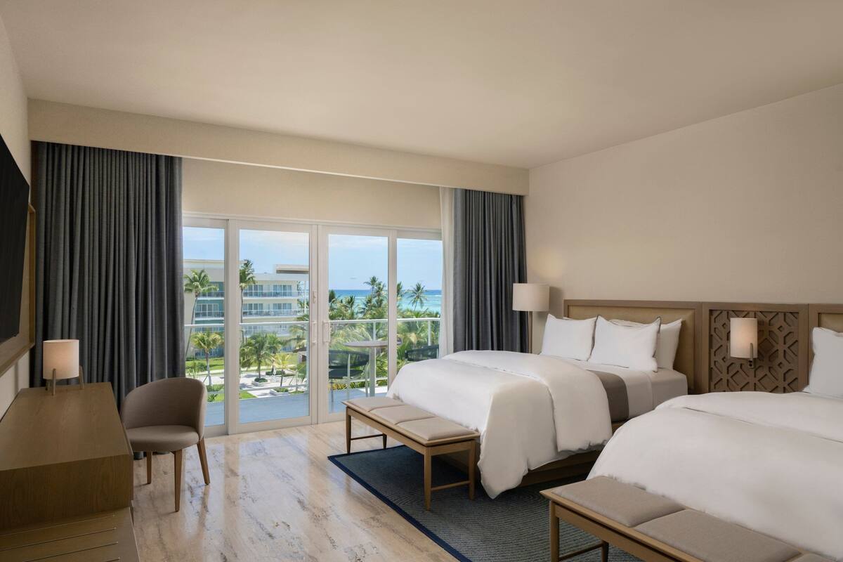 Une chambre d’hôtel avec deux lits, un bureau, une chaise et une vue sur l’océan et les palmiers.