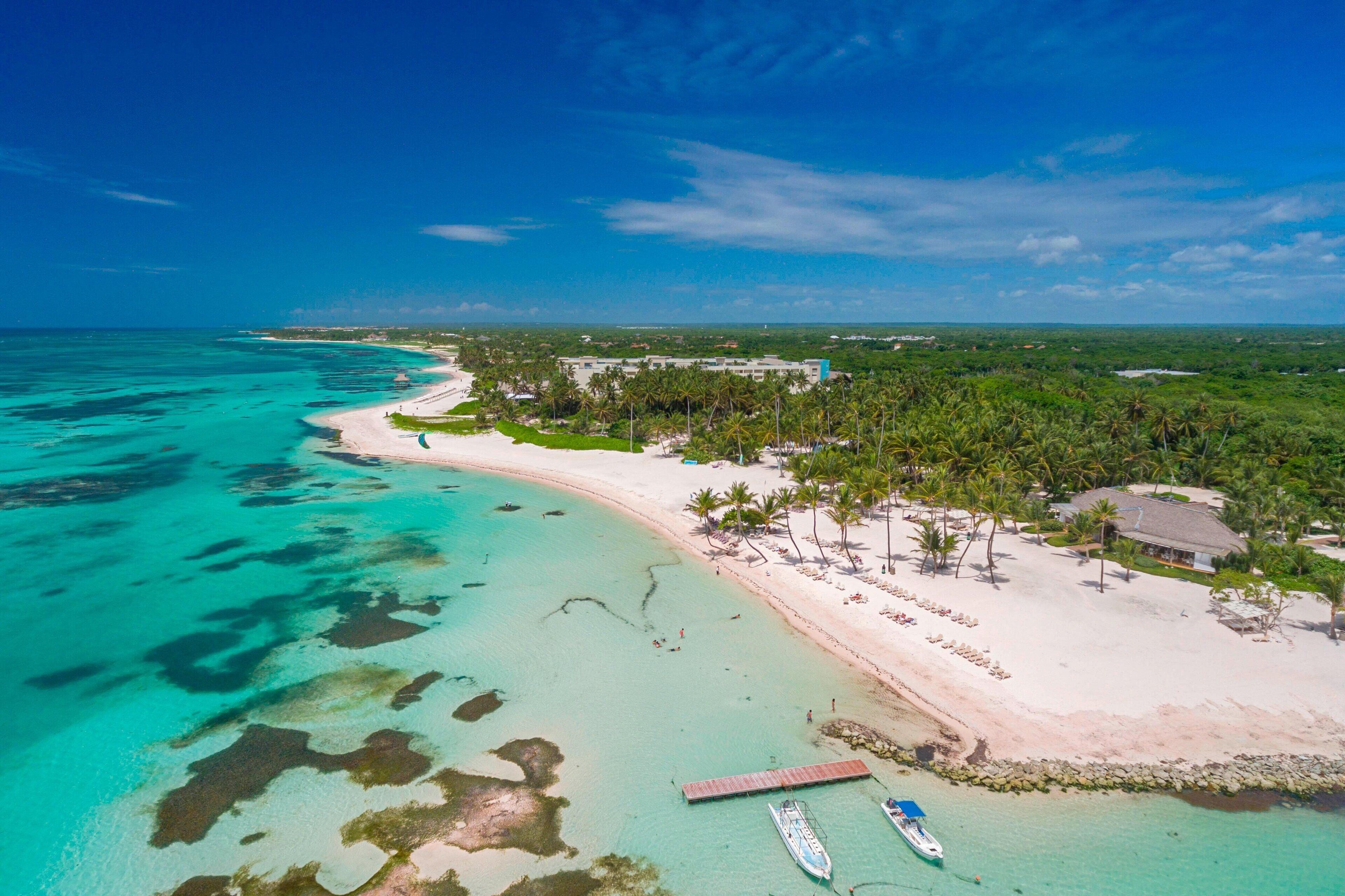 Une vue aérienne d’une plage aux eaux turquoise et limpides, bordée d’un rivage sablonneux et d’un petit quai où sont amarrés des bateaux.