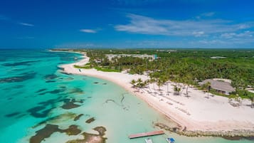 Une vue aérienne d’une plage aux eaux turquoise et limpides, bordée d’un rivage sablonneux et d’un petit quai où sont amarrés des bateaux.