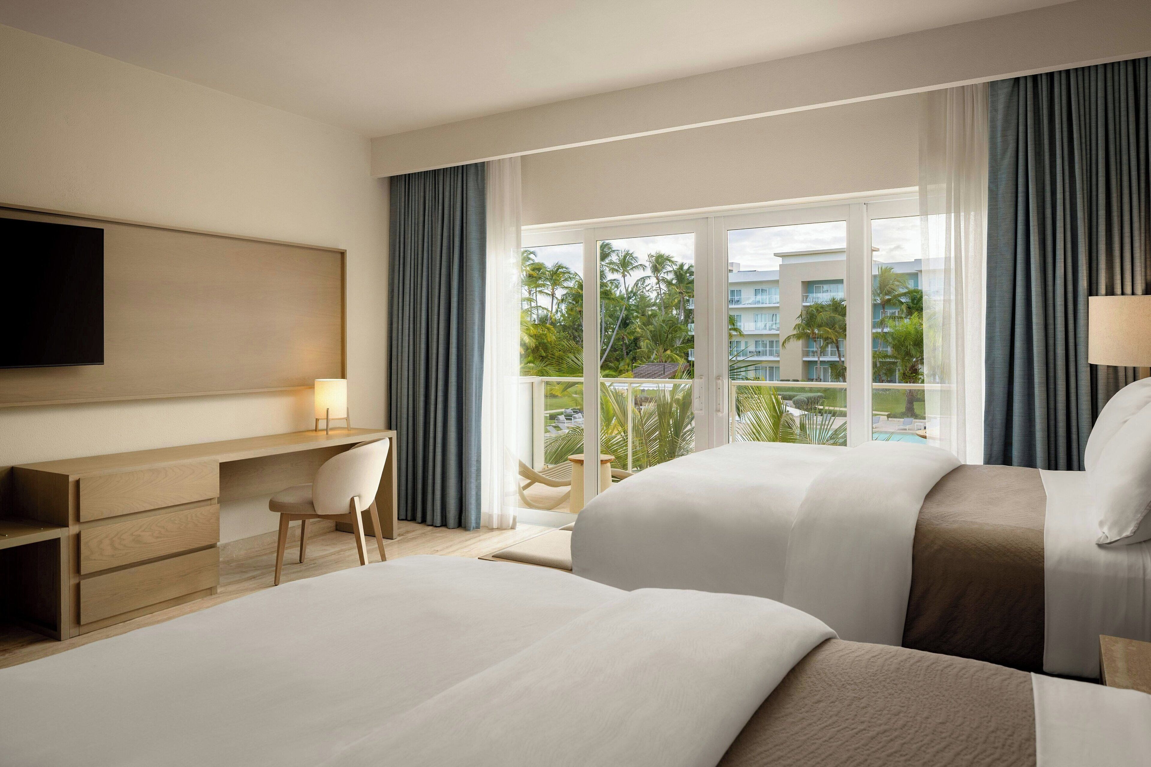 Une chambre d’hôtel avec deux lits, un bureau, une chaise et une grande fenêtre donnant sur des palmiers et des immeubles.