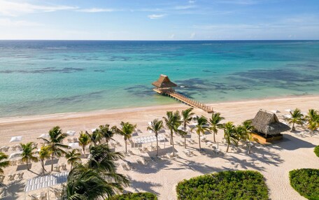 A beach resort with a thatched-roof structure, palm trees, and a pier extending into the turquoise sea.