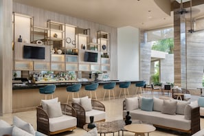 A modern bar area with a long counter, blue bar stools, and a variety of bottles. There are wicker chairs and a sofa with cushions.