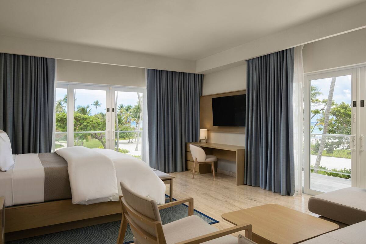 Une chambre d’hôtel avec un lit, un bureau, une chaise, une télévision et de grandes fenêtres avec des rideaux.