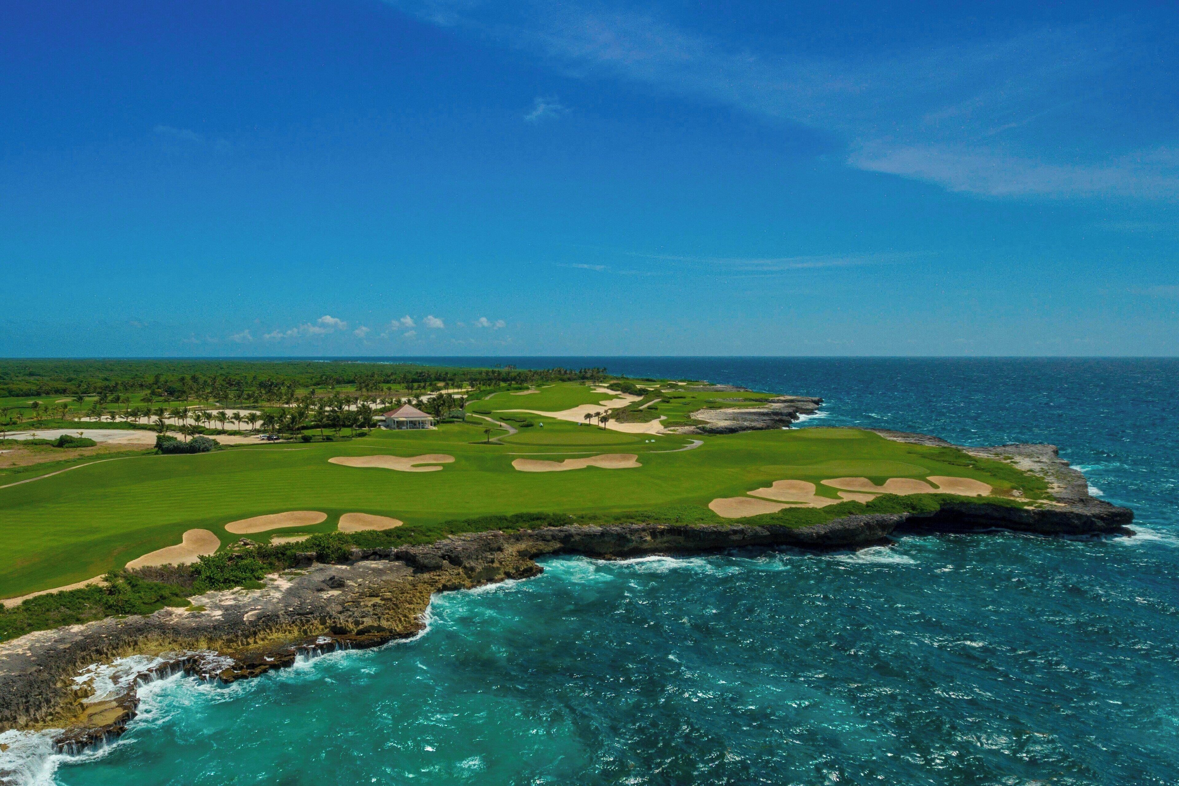 A coastal golf course with a clubhouse, green fairways, sand bunkers, and a rocky shoreline.