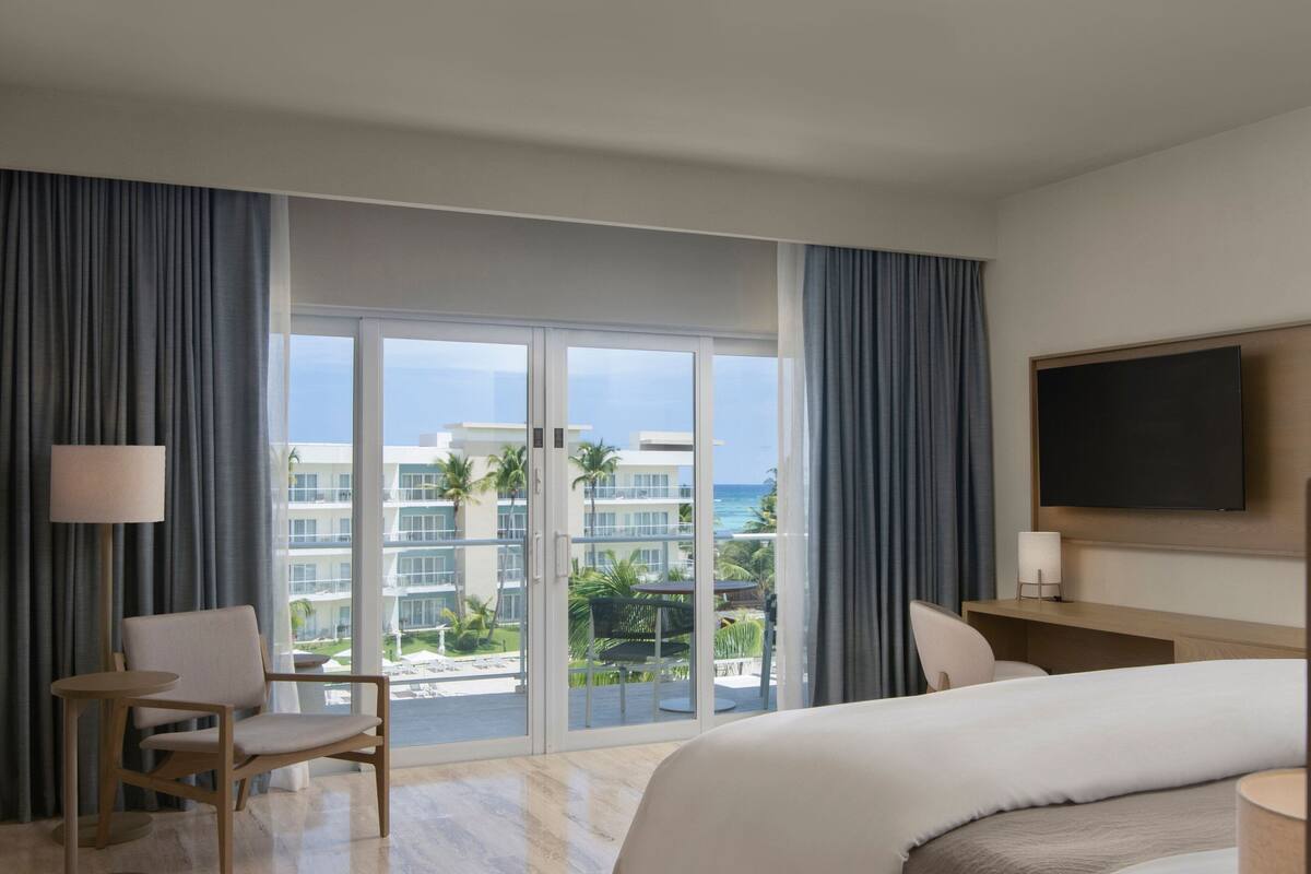Une chambre d’hôtel avec un grand lit, un bureau, une chaise et une télévision. Elle dispose d’un balcon avec vue sur l’océan et les bâtiments.