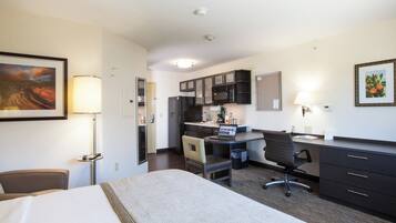 A hotel room with a bed, desk, chair, kitchenette, and a painting on the wall.