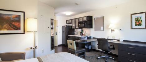 A hotel room with a bed, desk, chair, kitchenette, and a painting on the wall.
