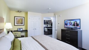 Habitación de hotel con cama, escritorio con una computadora portátil, televisión mostrando un noticiero y una kitchenette al fondo.