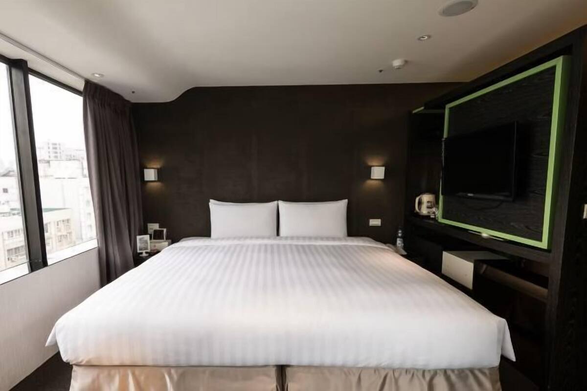 Habitación de hotel con una cama grande, televisión, mesita de noche y vistas a la ciudad a través de la ventana.