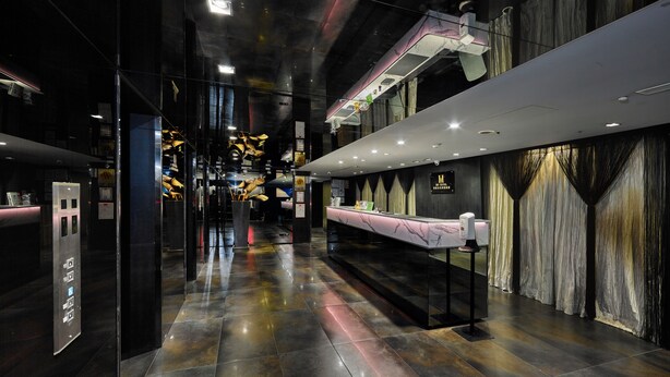 Un vestíbulo de hotel moderno con un mostrador de recepción de mármol, un ascensor de vidrio y un largo pasillo con un techo decorativo.
