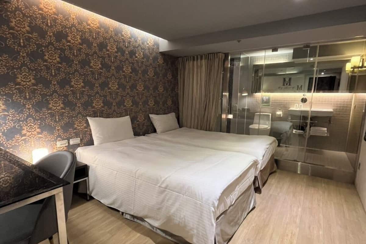 A modern hotel room with a large bed, a desk, a glass shower enclosure, and a patterned wallpaper.