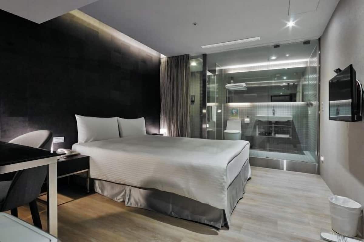A modern hotel room with a bed, a desk, a chair, a TV, and a glass partition separating the room from a bathroom.