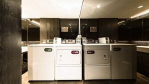 Una lavandería moderna con dos lavadoras y una secadora, equipada con controles digitales y señalización, en un ambiente limpio y con baldosas.