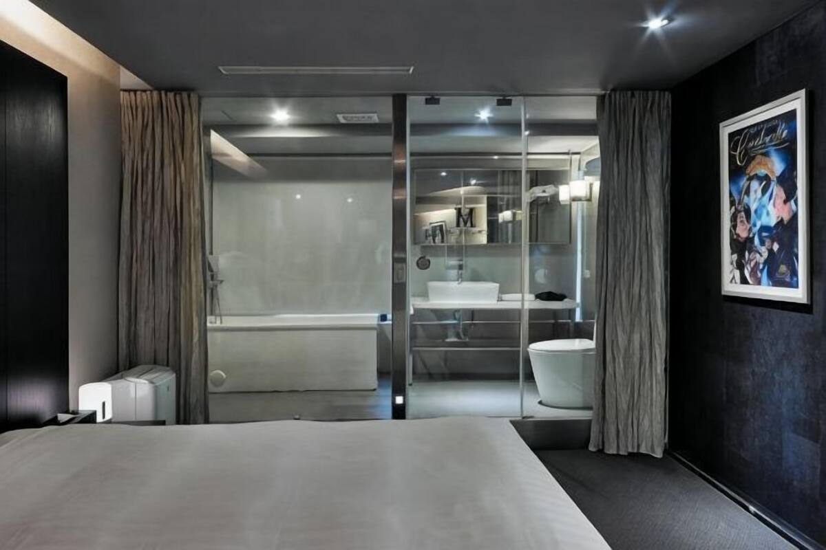 Una habitación de hotel moderna con una cama grande, zona de ducha con mampara de vidrio y baño con lavabo y espejo.