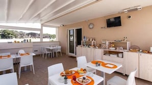 A dining area with white tables and chairs, a kitchen area with cabinets and a TV, and a view of the outdoors. - Bed & Breakfast Aria di Mare (Gallipoli)