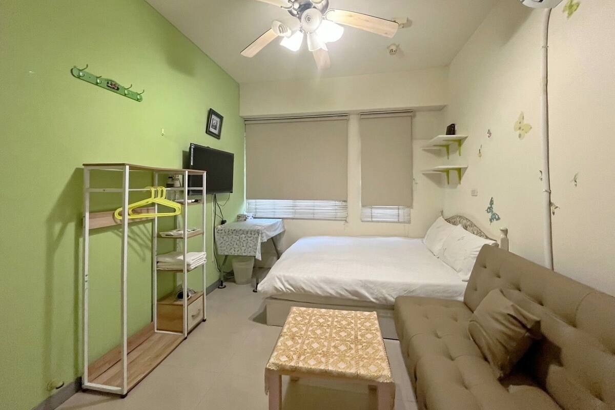 A bedroom with a bed, a sofa, a desk with a chair, a hanging light fixture, and a ceiling fan.