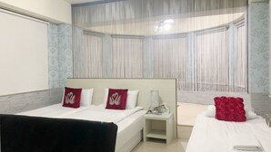 A hotel room with a large bed, a smaller bed, a desk, and a chair. There is a window with sheer curtains and a ceiling light.