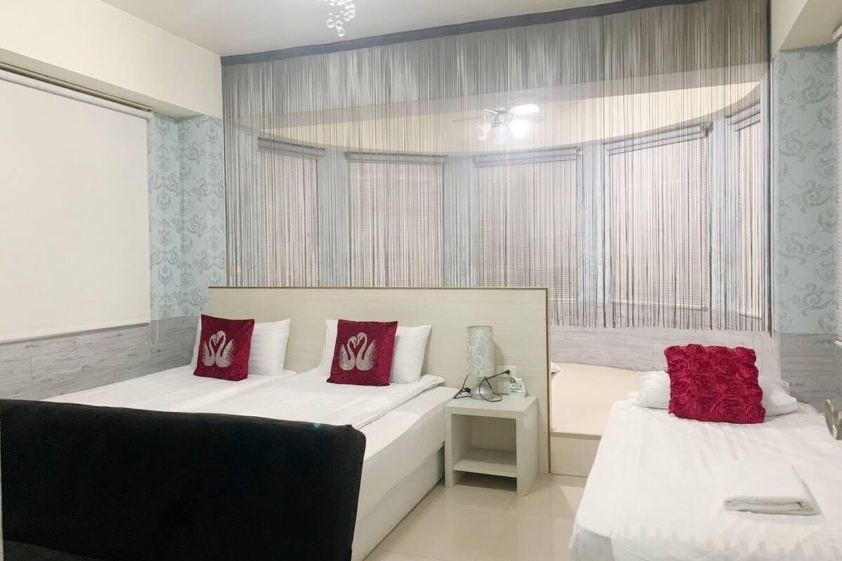 A hotel room with a large bed, a smaller bed, a desk, and a chair. There is a window with sheer curtains and a ceiling light.