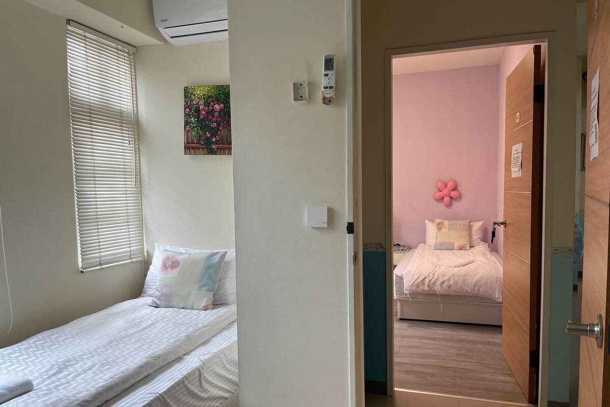 A bedroom with a single bed, a window with blinds, a wall-mounted air conditioner, and a door leading to another room.