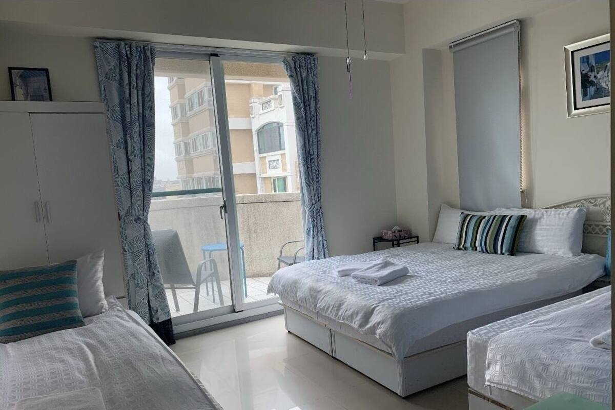 A hotel room with a large bed, a smaller bed, a wardrobe, a desk, and a balcony with a view of the building.
