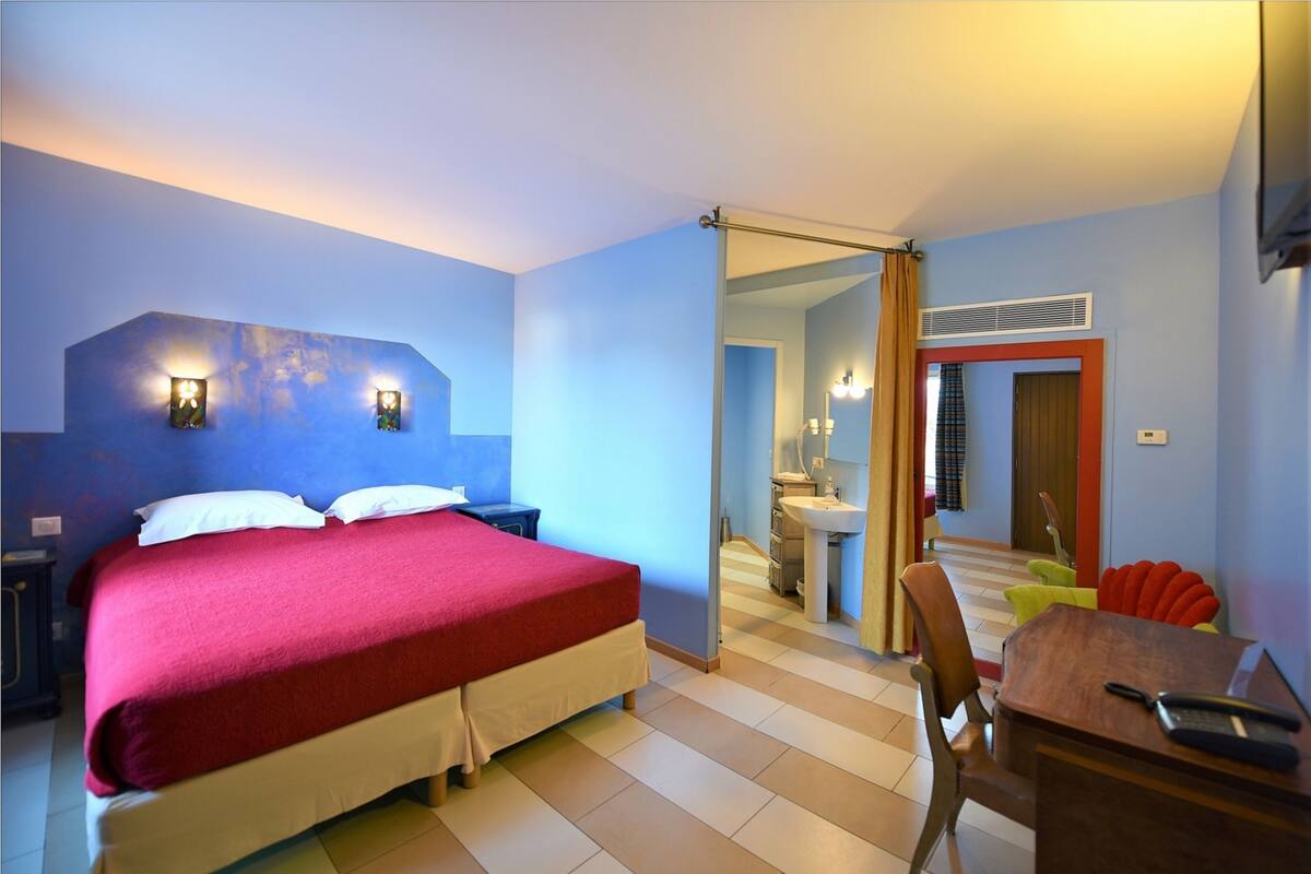 A hotel room with a bed, bedside tables, a desk, a chair, a mirror, a sink, and a shower area.