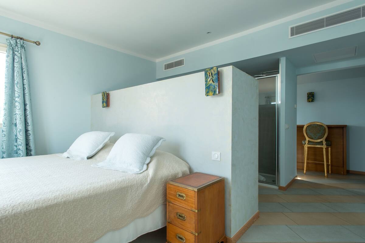 A bedroom with a bed, a wooden chest of drawers, a chair, and a bathroom visible through a glass door.