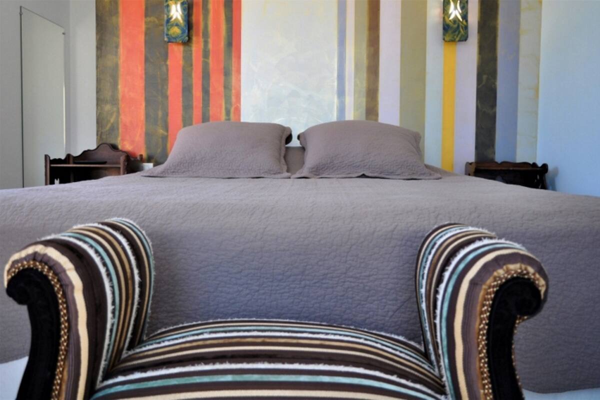 A bed with two pillows, a striped armchair, and a wall with vertical stripes and two wall sconces.