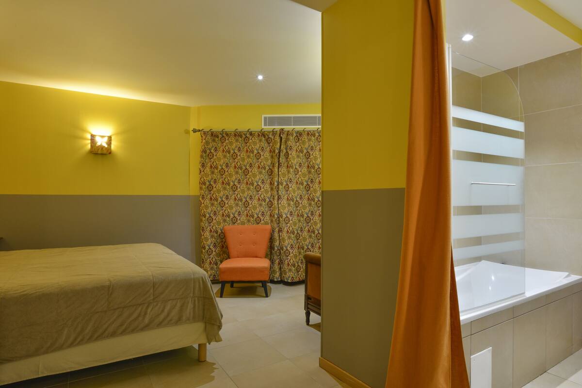 A hotel room with a bed, a chair, a shower area, and a curtain.